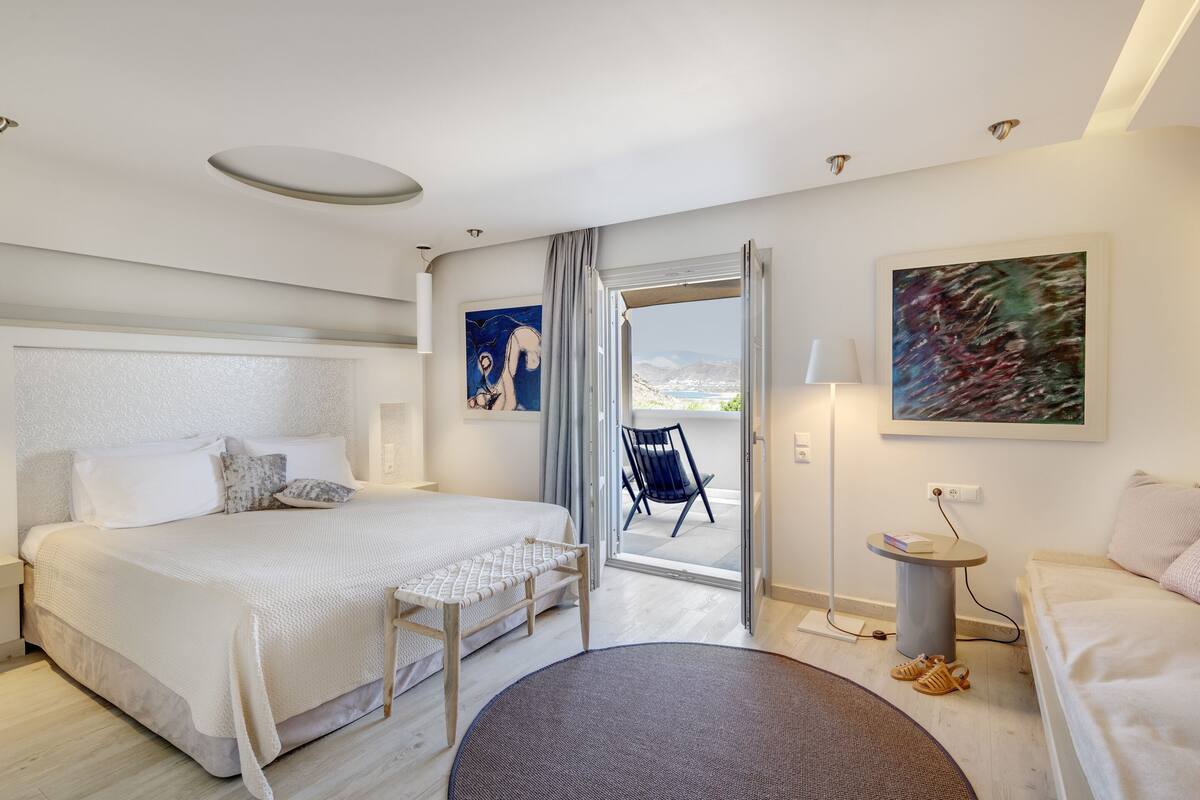 Ein modernes Hotelzimmer mit einem großen Bett, einem kleinen Schreibtisch, einem Sessel und Blick ins Freie.