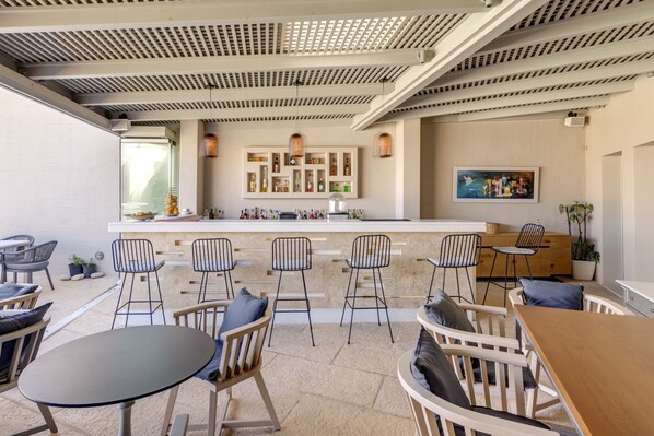An outdoor seating area with tables and chairs, a bar with shelves of bottles, and a wall with framed artwork. - Kouros Art Hotel - Adults Only (Naxos)