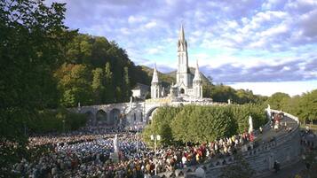 A large outdoor amphitheater with a crowd of people, surrounded by trees and a historic building with two towers.