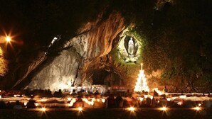 Uma gruta com uma estátua iluminada por luzes, cercada por uma multidão de pessoas e velas.