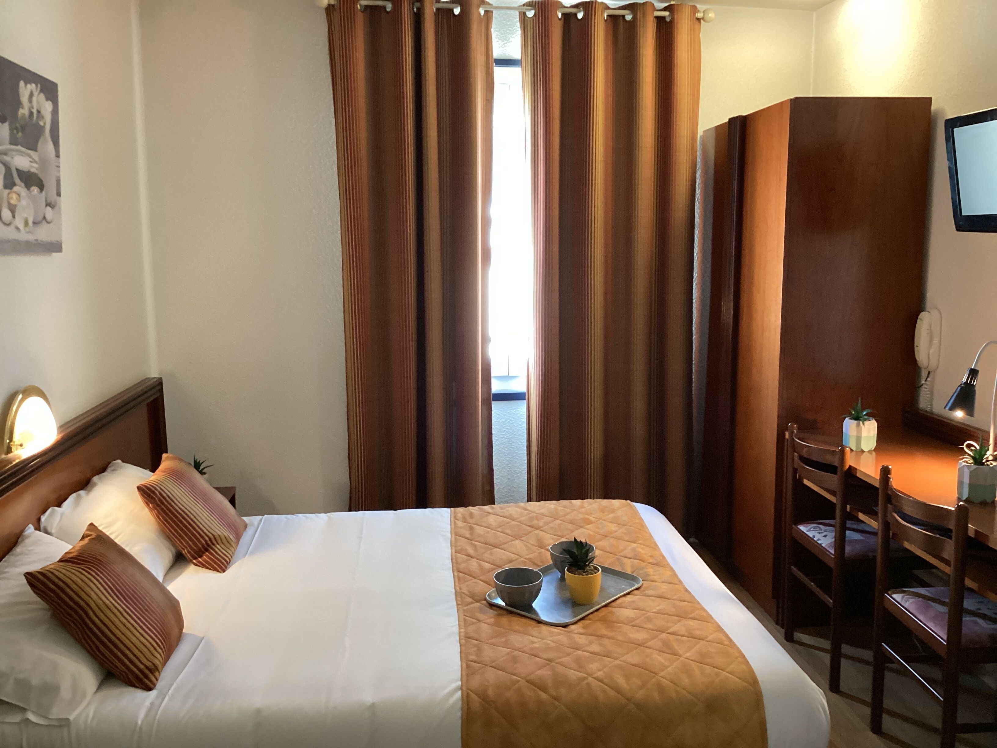 Ett hotellrum med en säng, ett skrivbord, en stol, ett litet bord med en växt och ett fönster med gardiner.
