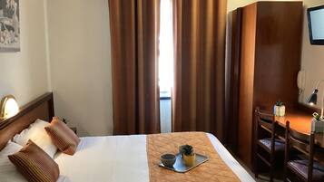 Ett hotellrum med en säng, ett skrivbord, en stol, ett litet bord med en växt och ett fönster med gardiner.