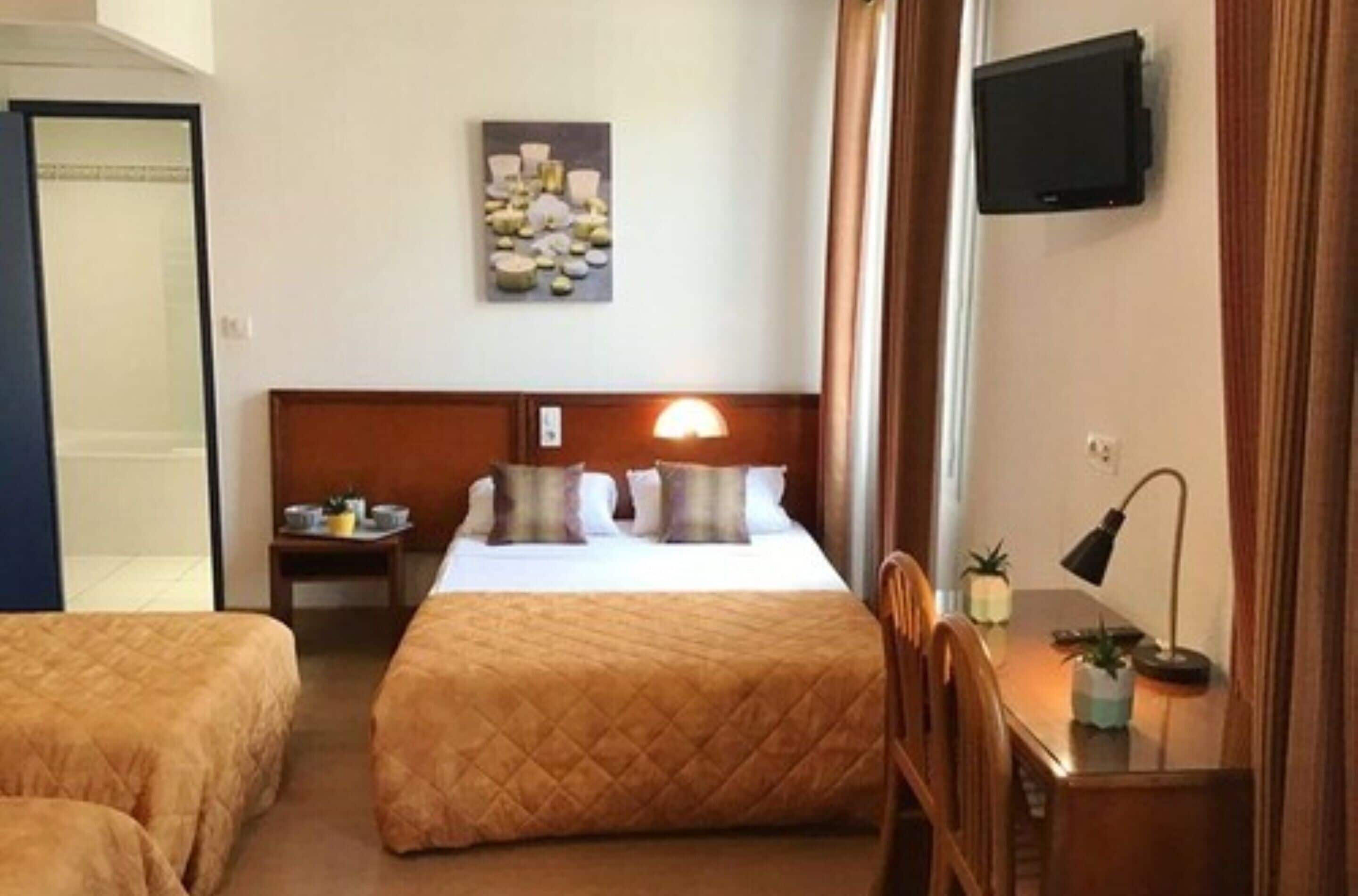 A hotel room with two beds, a bedside table with a lamp, a TV mounted on the wall, and a painting above the bed.