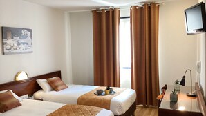 Quarto de hotel com duas camas, uma escrivaninha com luminária, televisão e janela com cortinas.