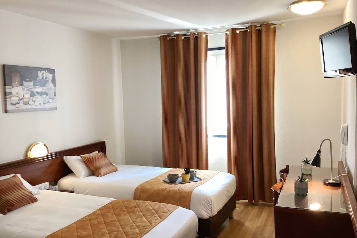Habitación de hotel con dos camas, un escritorio con lámpara, televisor y ventana con cortinas.