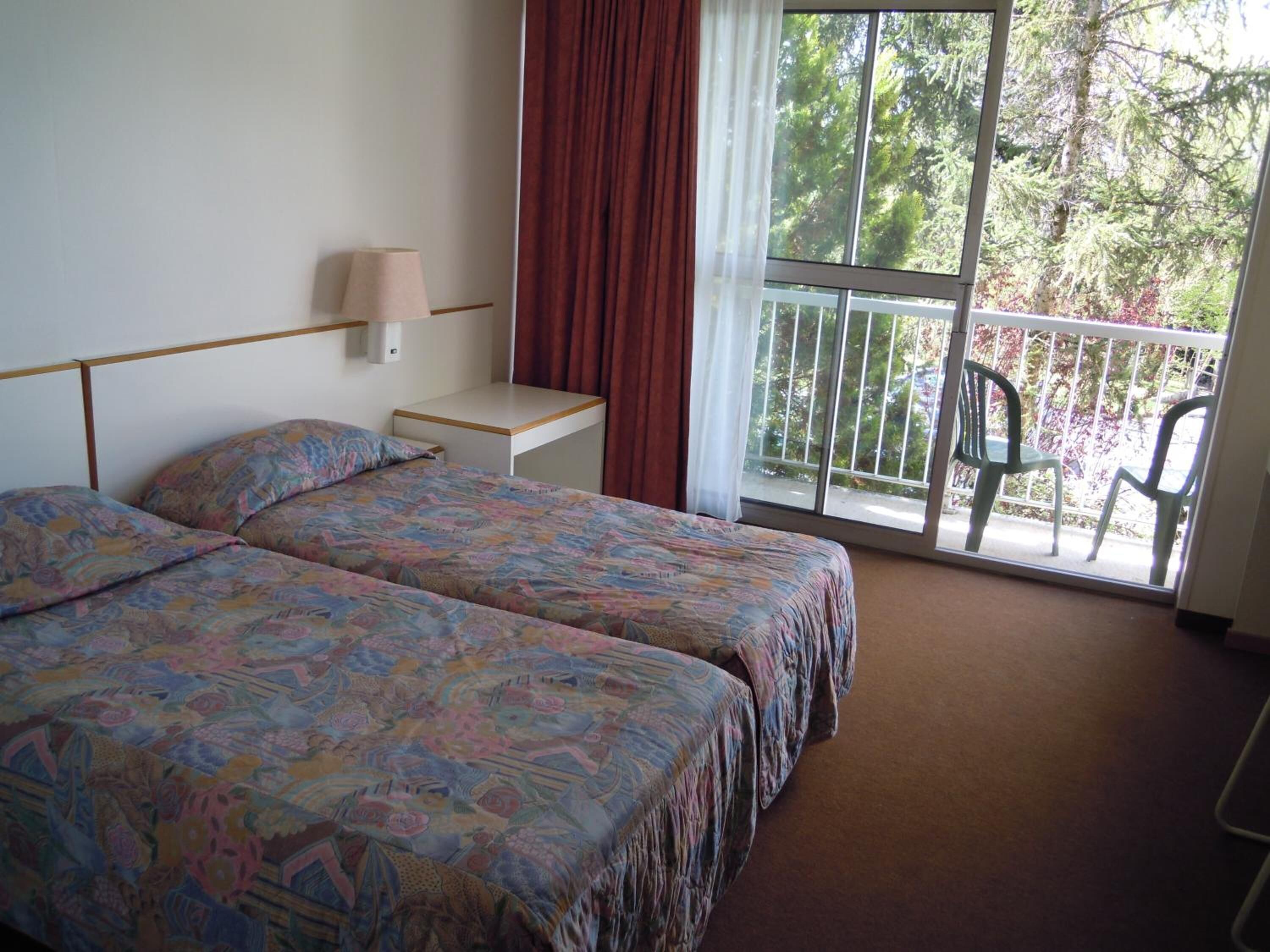 A hotel room with two beds, a nightstand, a lamp, a window with a view of trees, and a balcony with chairs.