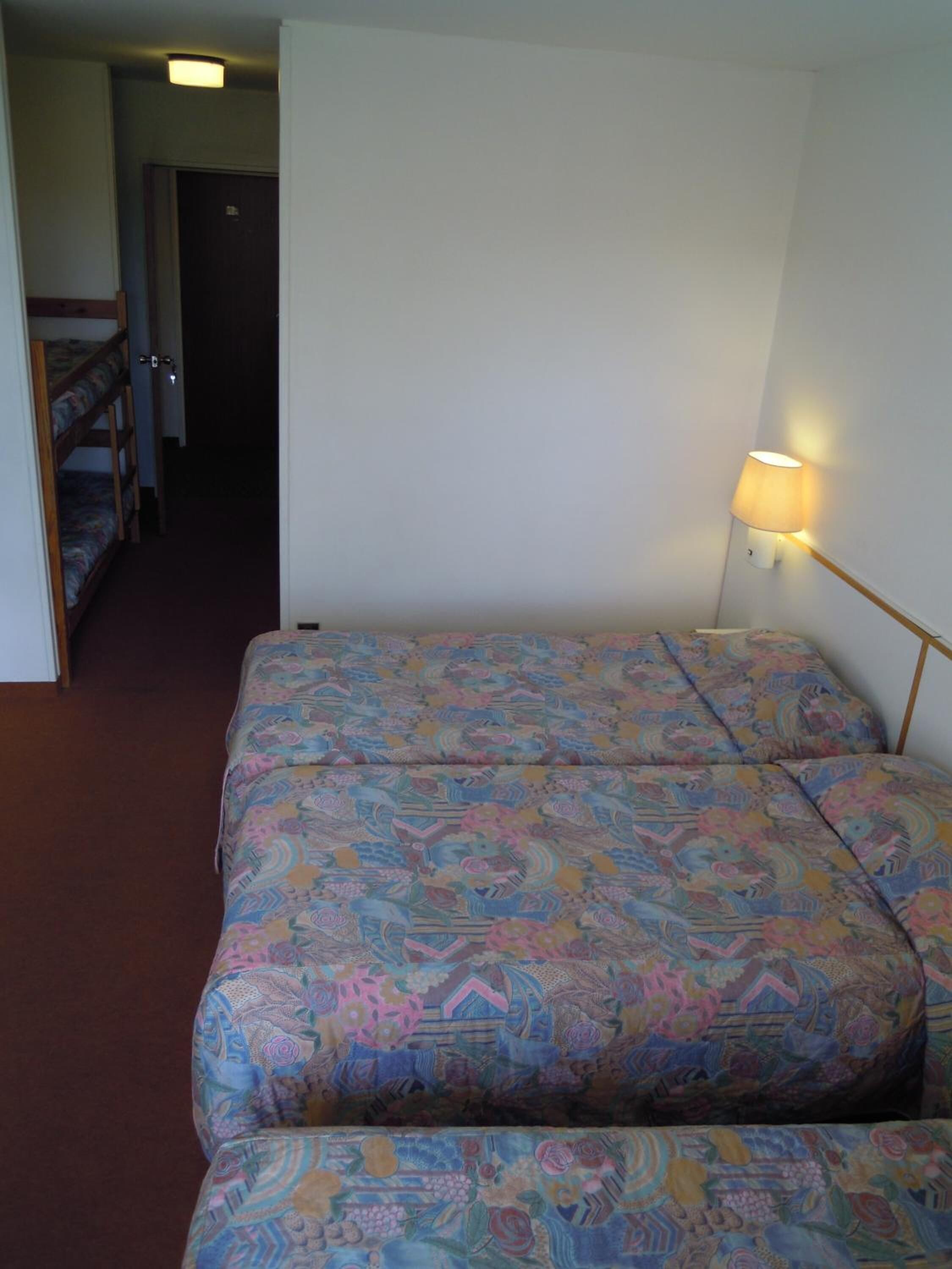Una habitación con una cama individual superpuesta, una cama sencilla con ropa de cama estampada, una lámpara de noche y una puerta que da a otra habitación.