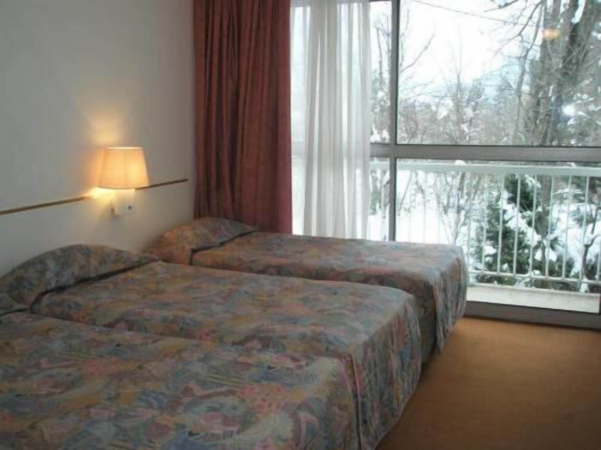 Habitación de hotel con dos camas, una ventana que da a árboles y una lámpara fijada a la pared.
