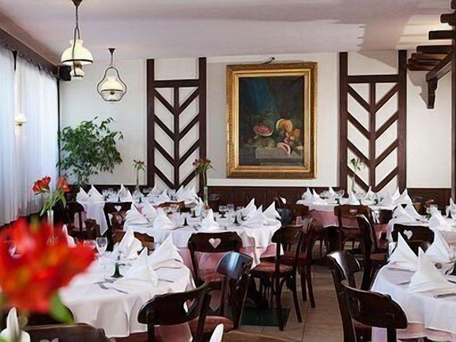 Een restaurant met meerdere tafels, bedekt met witte tafelkleden, houten stoelen en hangende lampen.