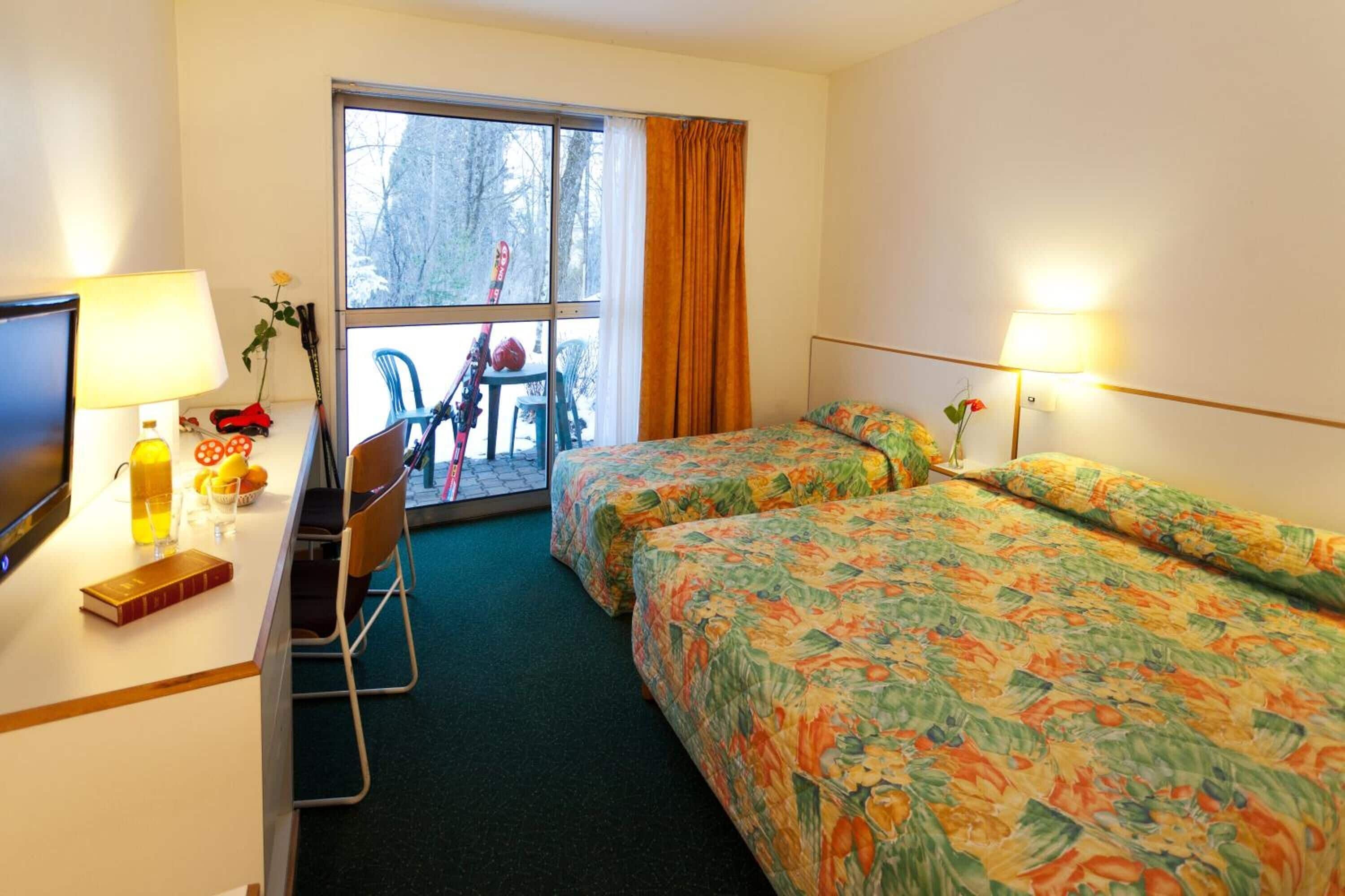 Una habitación de hotel con dos camas, un escritorio con televisor, una silla, una ventana con cortinas y vistas a árboles.