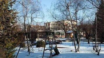 A snow-covered outdoor area with a swing set, a building in the background, and leafless trees.
