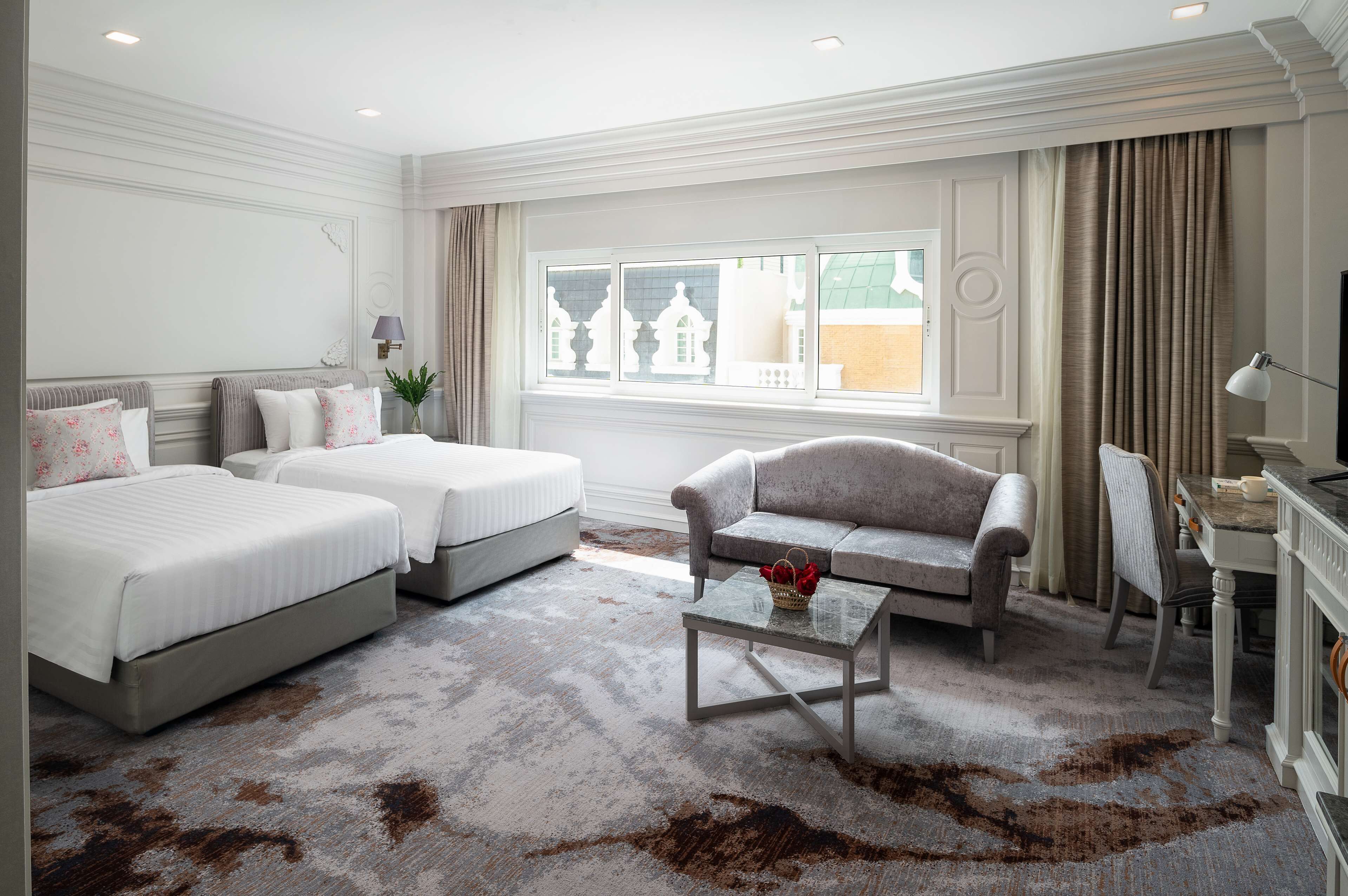 A hotel room with two beds, a sofa, a small table, a TV, and a window with a view of buildings.