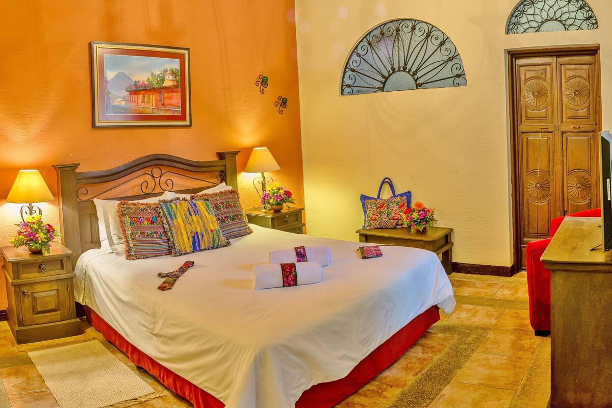 Une chambre d’hôtel élégamment aménagée, dotée d’un grand lit, de deux tables de chevet avec des lampes, d’un mur d’accent rouge, d’un ventilateur décoratif et d’une armoire en bois.