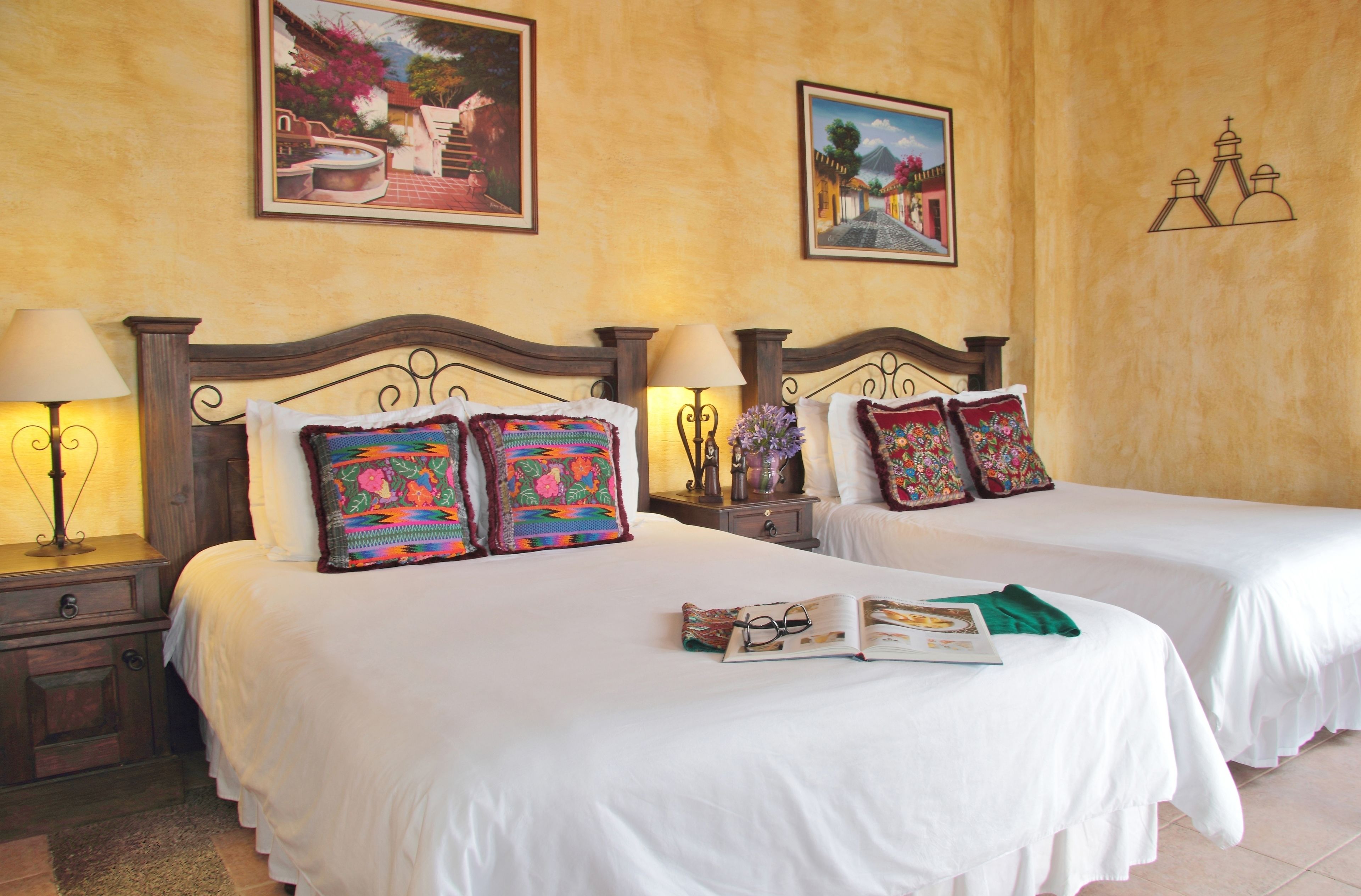 A hotel room with two beds, each with a white bedspread and colorful pillows. There are two bedside tables with lamps, a nightstand, and a wall-mounted shelf with decorative items.