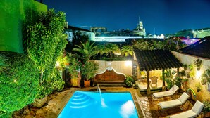 A swimming pool with lounge chairs, surrounded by greenery and a view of a historic building at night. - Hotel Casa del Parque (Antigua Guatemala)