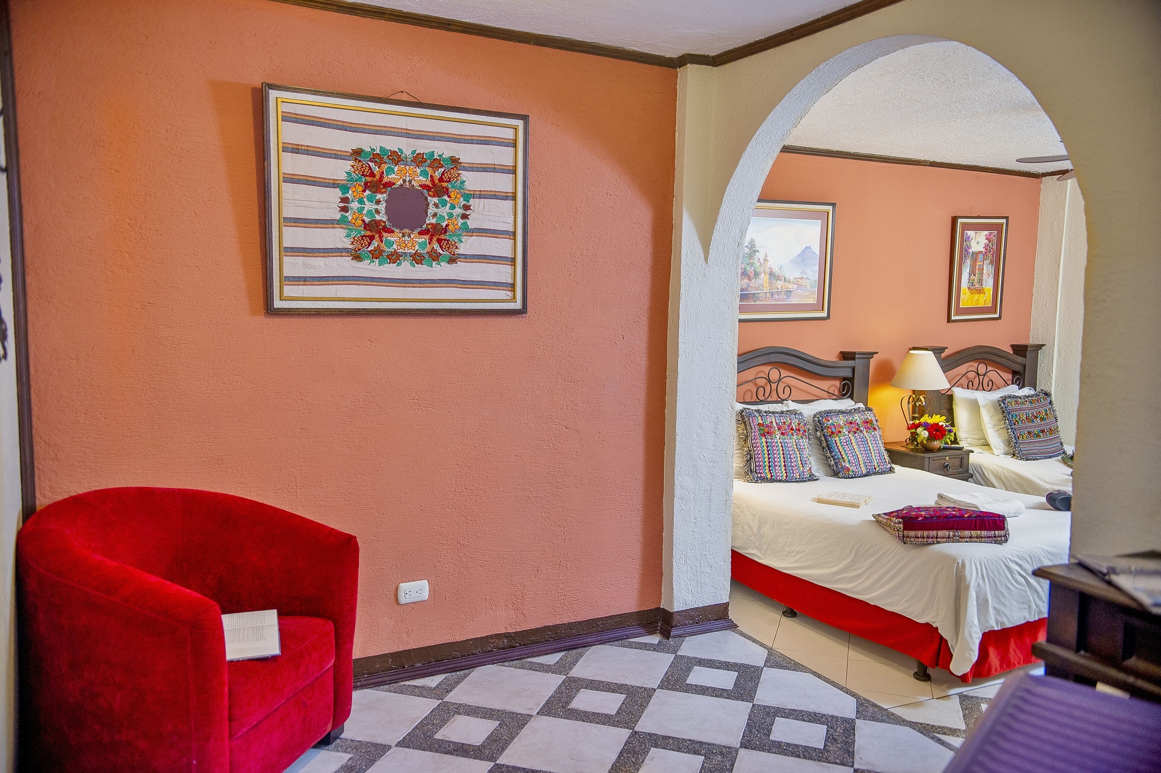 Une chambre d’hôtel avec un fauteuil rouge, un lit doté de plusieurs oreillers, une table de chevet avec une lampe et une œuvre d’art encadrée au mur.