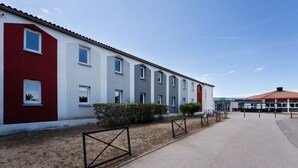 A modern building with a red and white facade, multiple windows, and a paved entrance area. - Kyriad ECO - Perpignan Nord Aéroport (Rivesaltes)