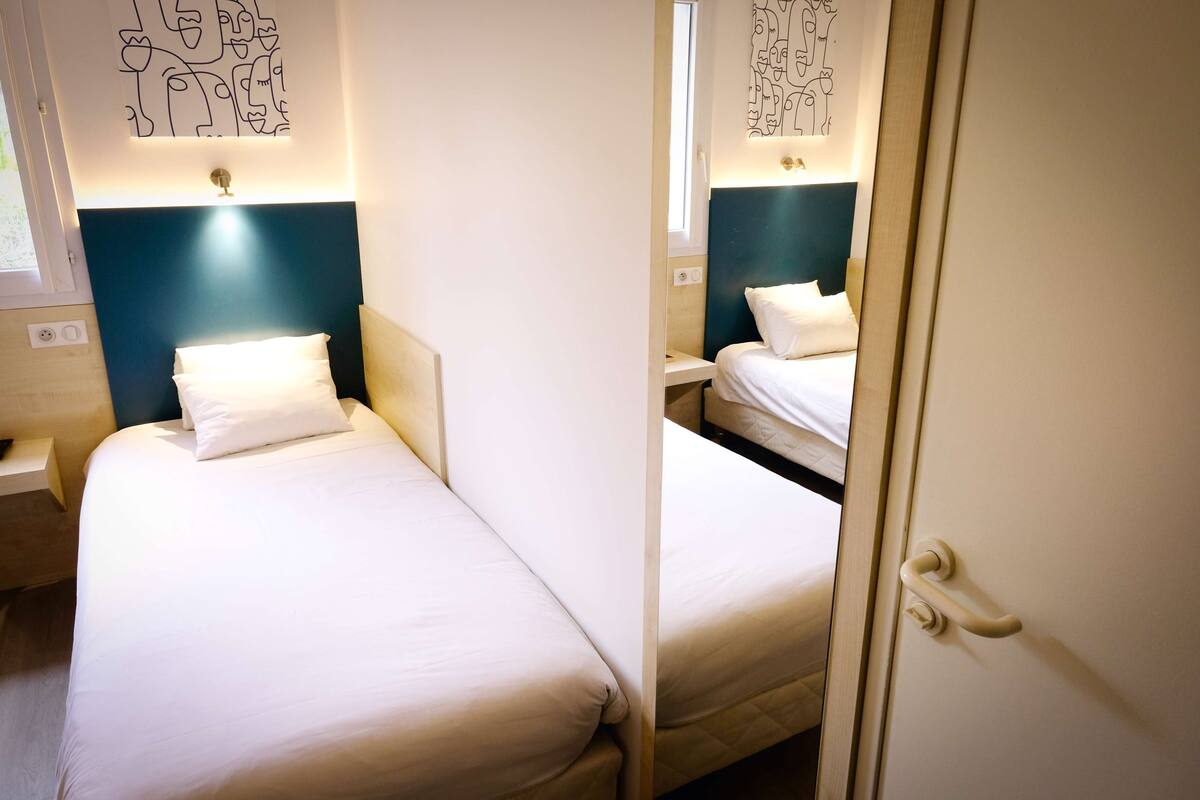 A hotel room with two beds, a nightstand, a wall-mounted light, and a window.