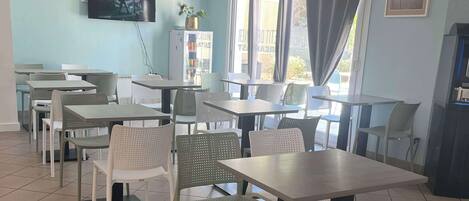 A dining area with multiple tables and chairs, a television mounted on the wall, and large windows with curtains.