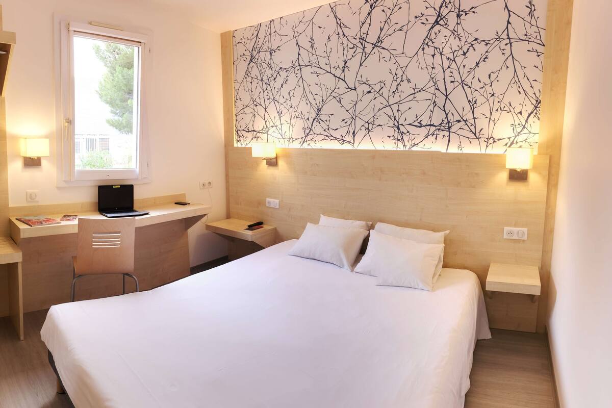 A hotel room with a bed, a desk with a laptop, a chair, a nightstand, and a wall with a branch pattern.