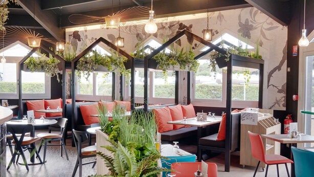A restaurant interior with booths, tables, hanging plants, and a view of the outdoors. - Kyriad ECO - Perpignan Nord Aéroport (Rivesaltes)