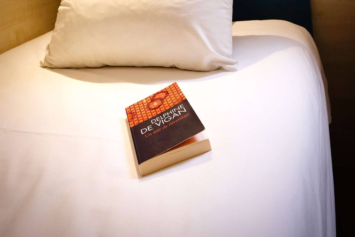 A neatly made bed with white bedding and pillows, a book titled 'Delphine De Vigan' on the bed, and a wall-mounted light fixture above.