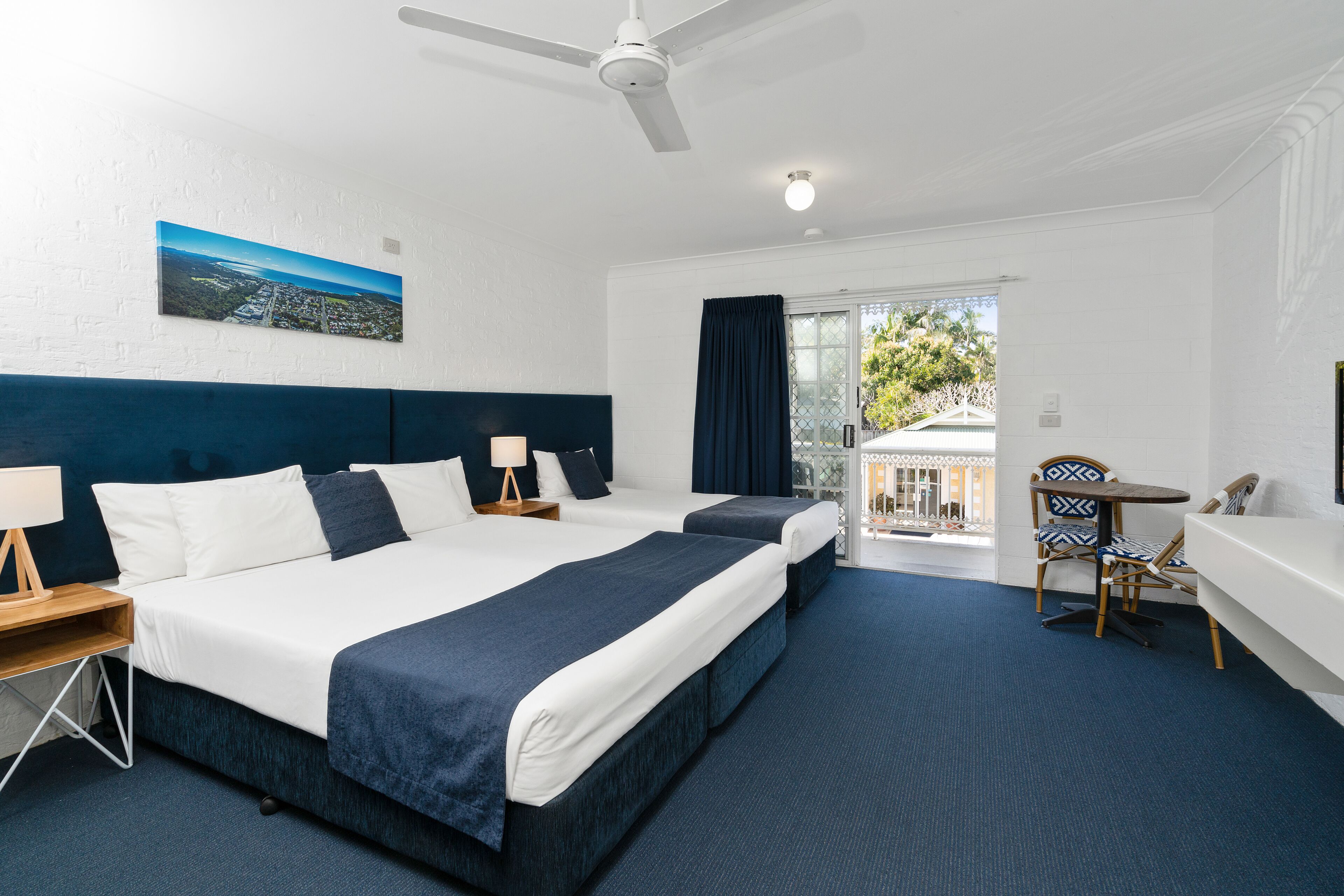 A hotel room with two beds, a desk, a chair, a balcony, and a ceiling fan.