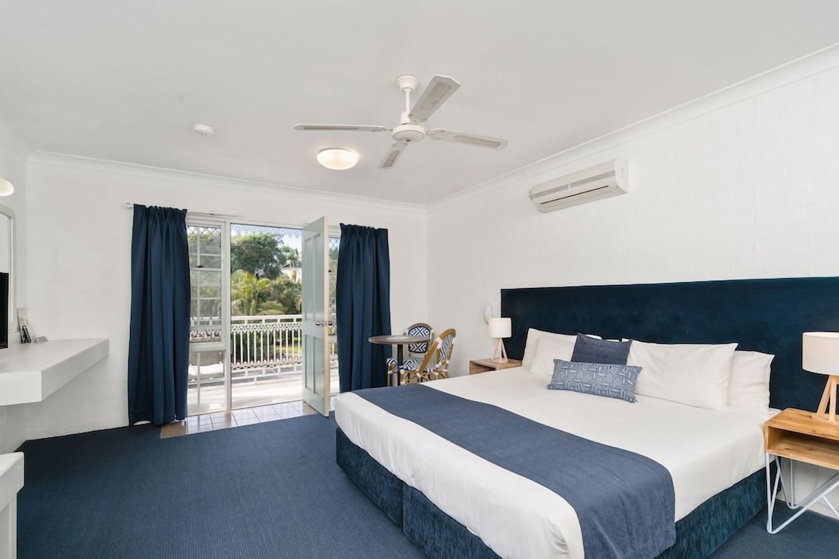 A hotel room with a bed, a ceiling fan, a window with curtains, a small table with a chair, and a balcony.