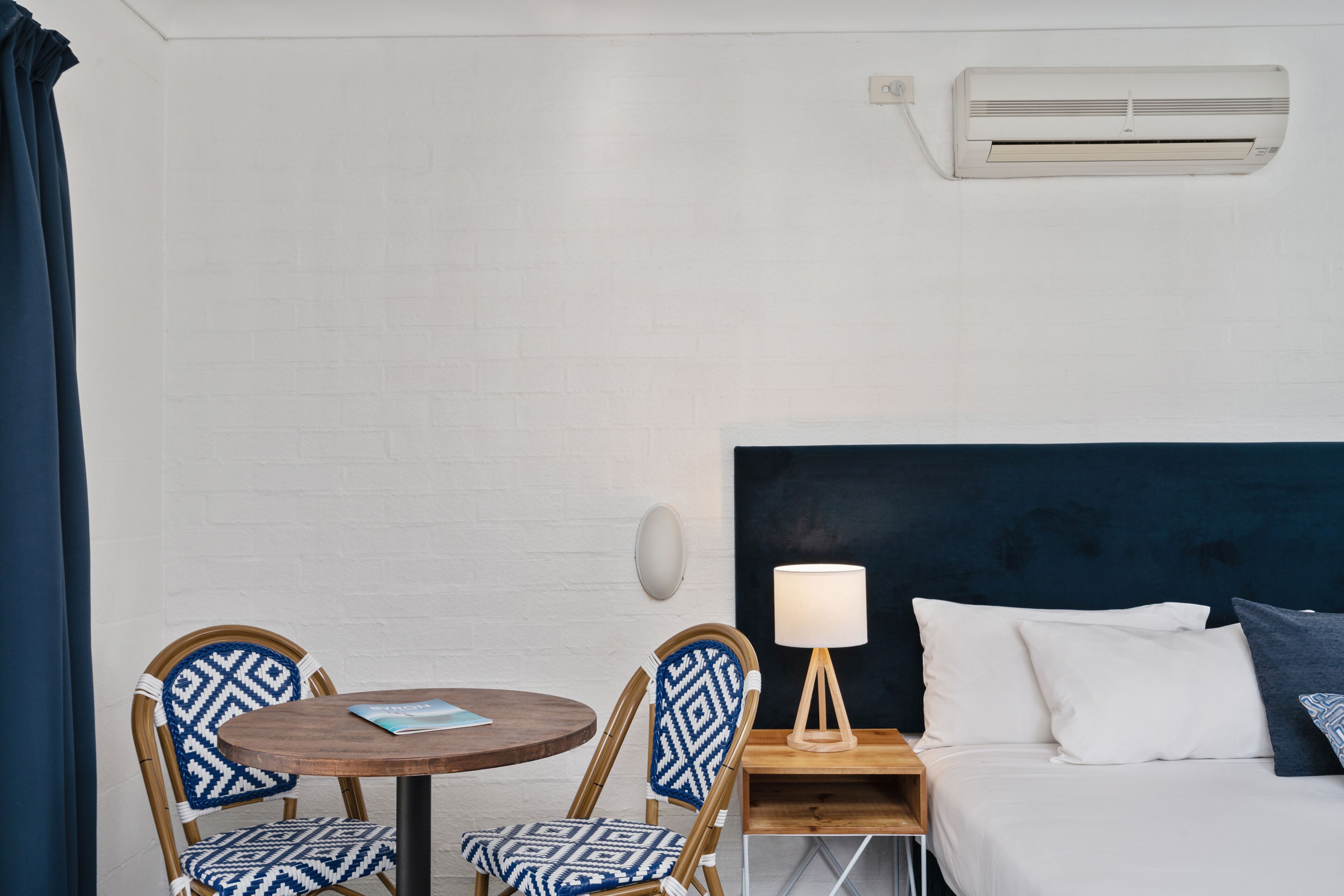 A hotel room with a bed, two patterned chairs, a small table, a lamp, and an air conditioning unit.