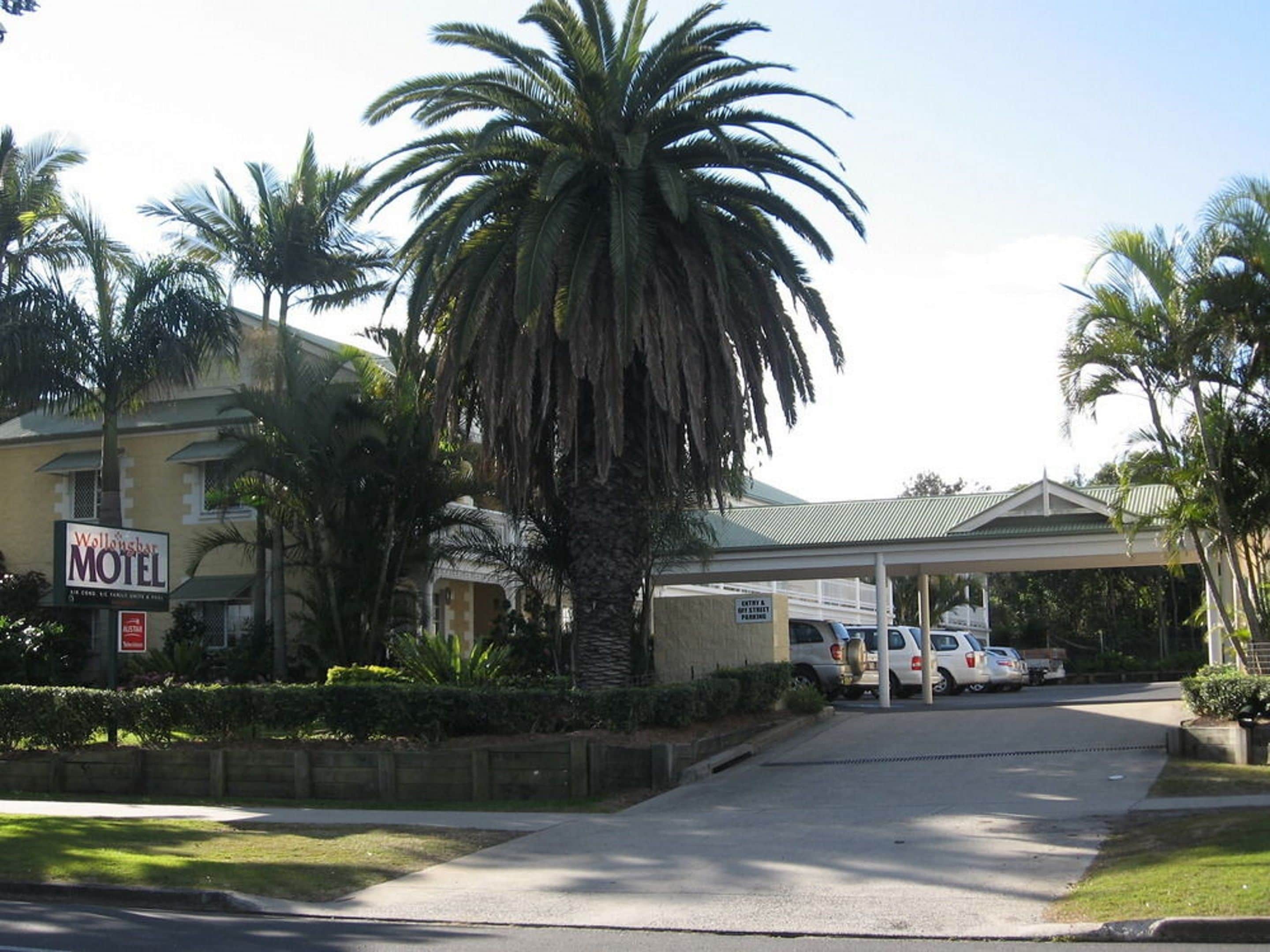 Ein Motel mit überdachtem Parkbereich, Palmen und einem Schild mit der Aufschrift 'Motel' und 'Parken'.