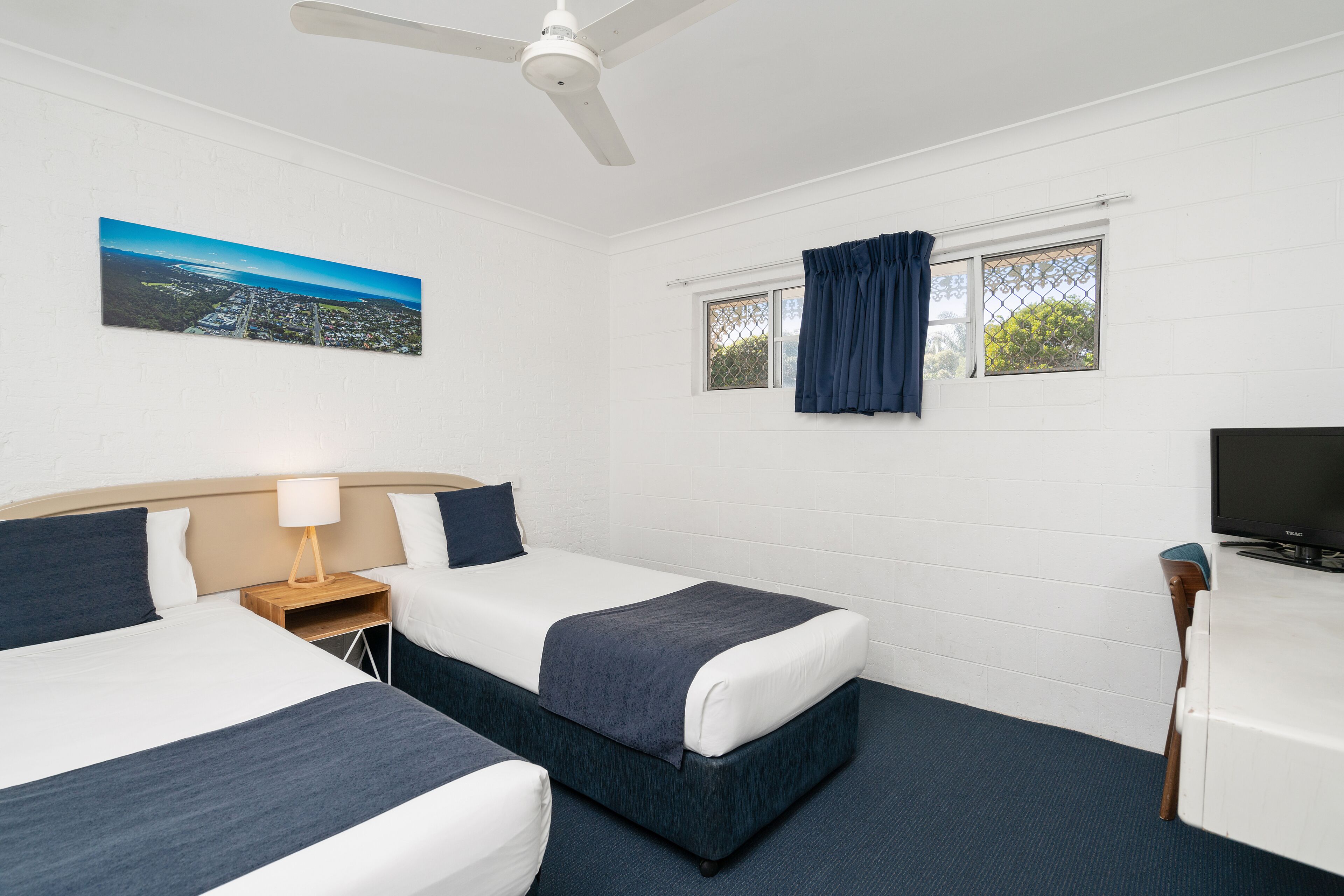 Une chambre d’hôtel avec deux lits, un ventilateur de plafond, une fenêtre avec des rideaux, une télévision et un tableau au mur.