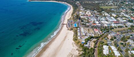 An aerial view of a coastal town with a beach, residential areas, and a lighthouse on a hill.