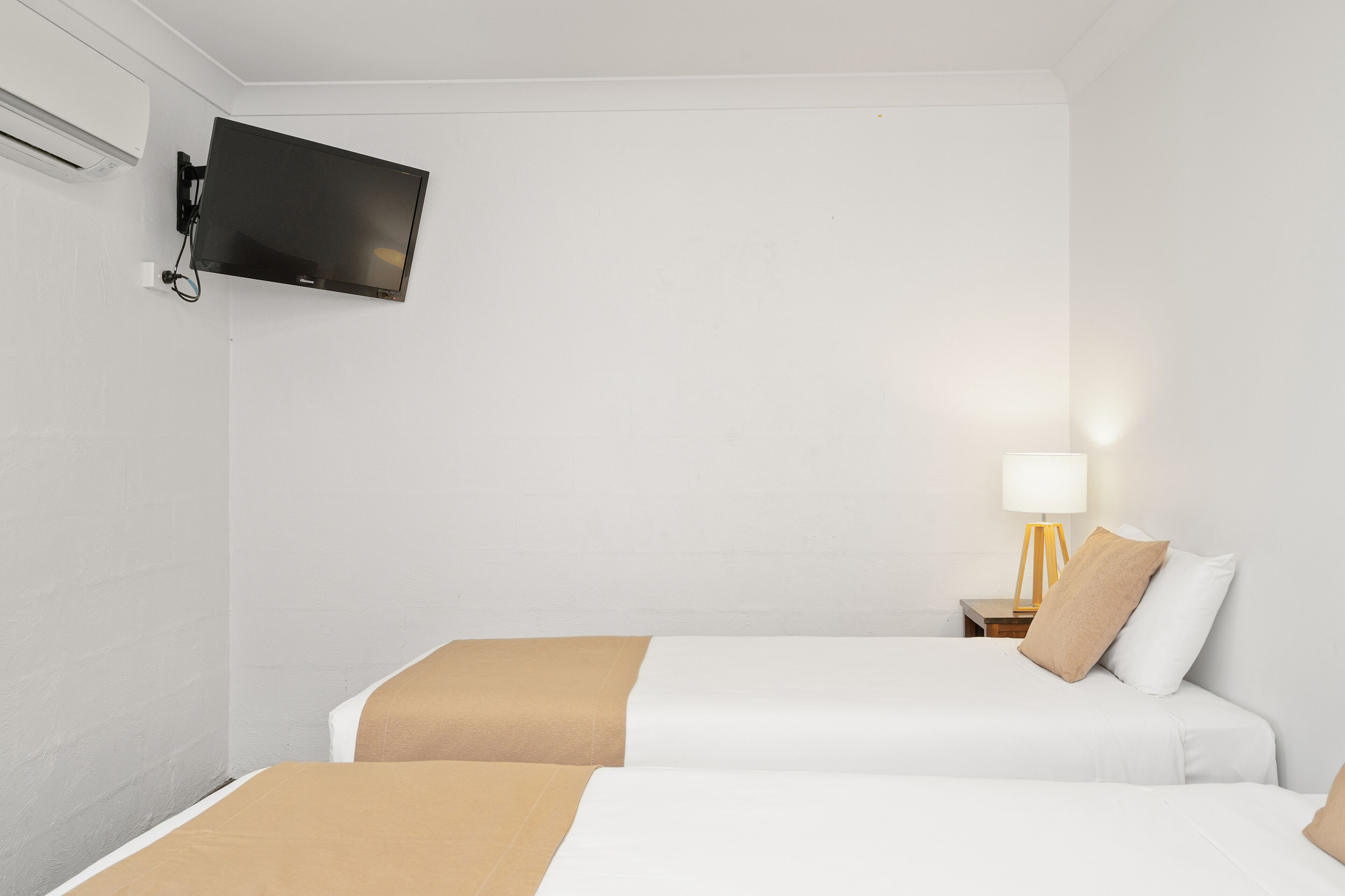 A hotel room with two beds, a flat-screen TV mounted on the wall, a bedside table with a lamp, and a wall-mounted air conditioner.