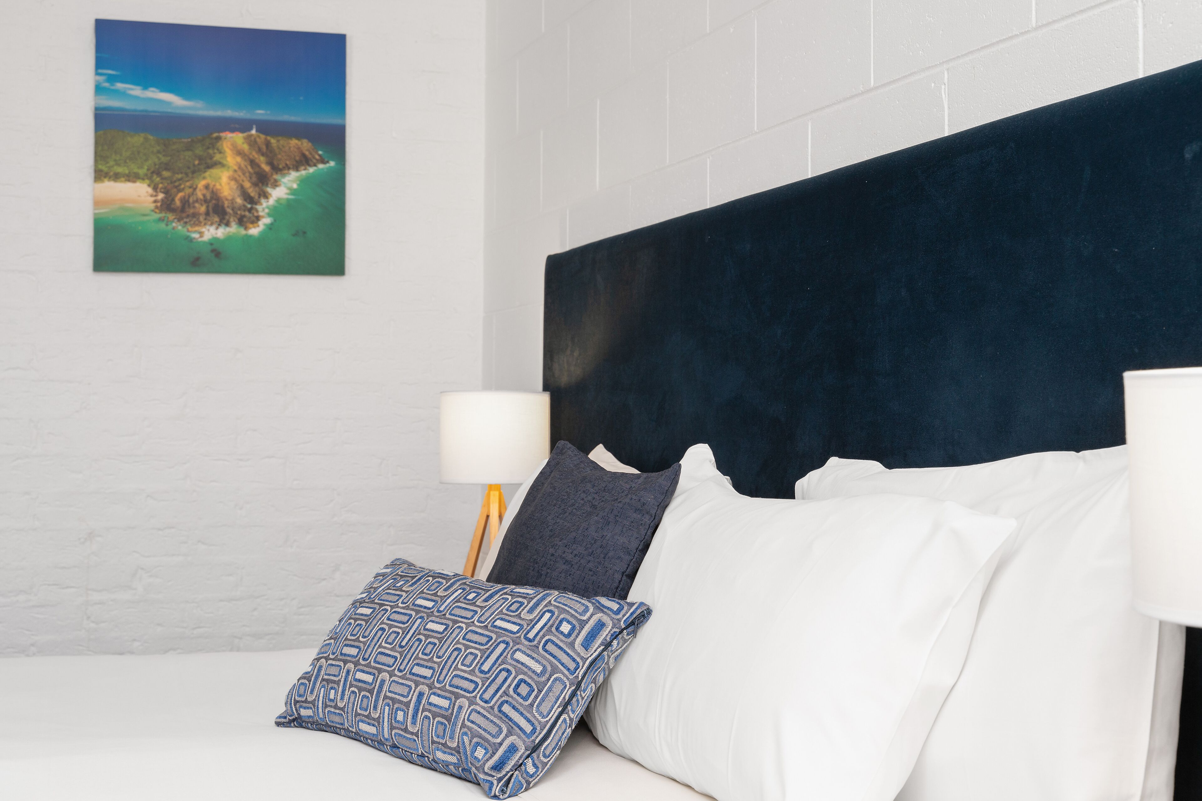 A hotel room with a white brick wall, a dark headboard, white bedding, and a patterned pillow.