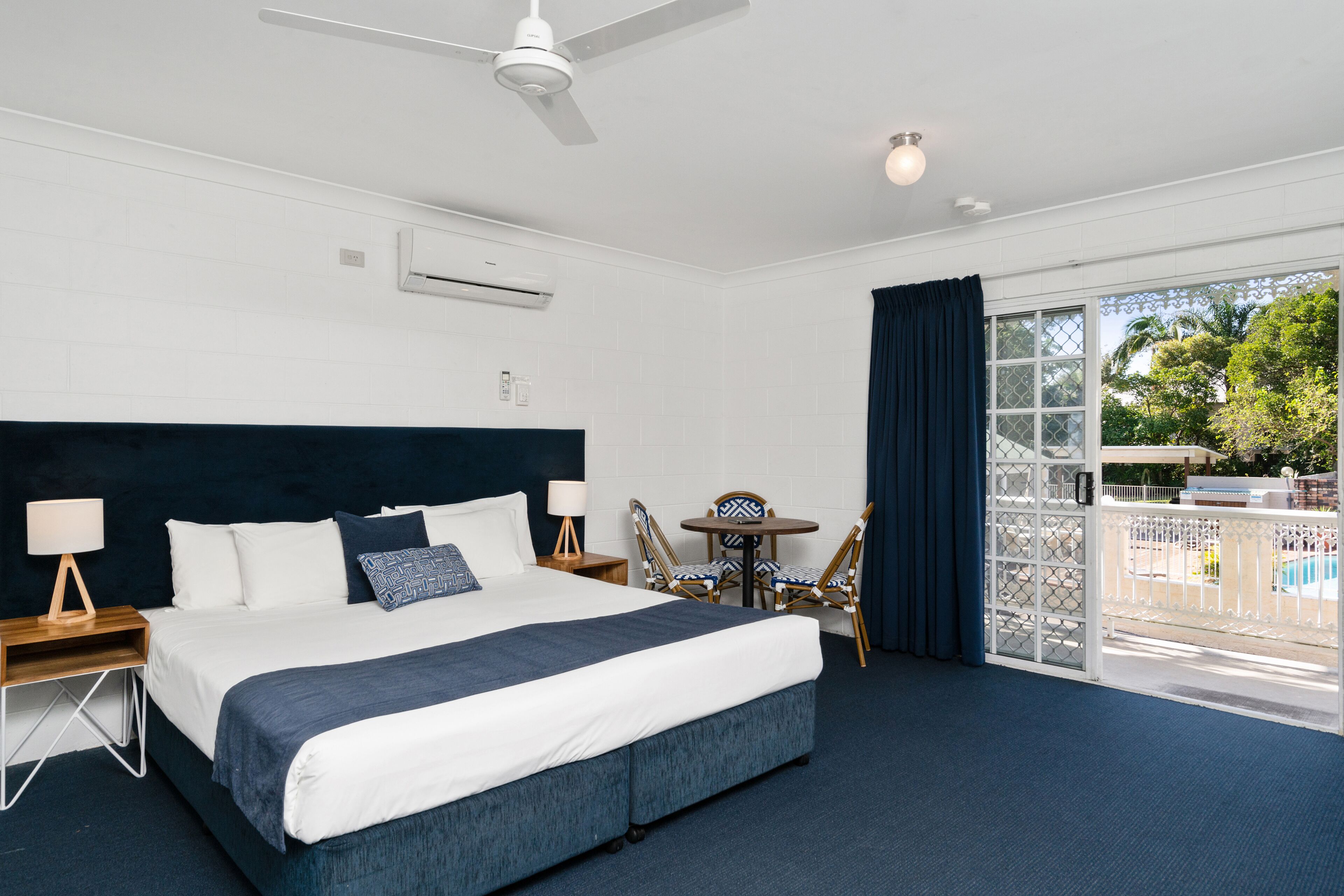A hotel room with a large bed, a ceiling fan, a window with curtains, a small table with chairs, and an air conditioning unit.