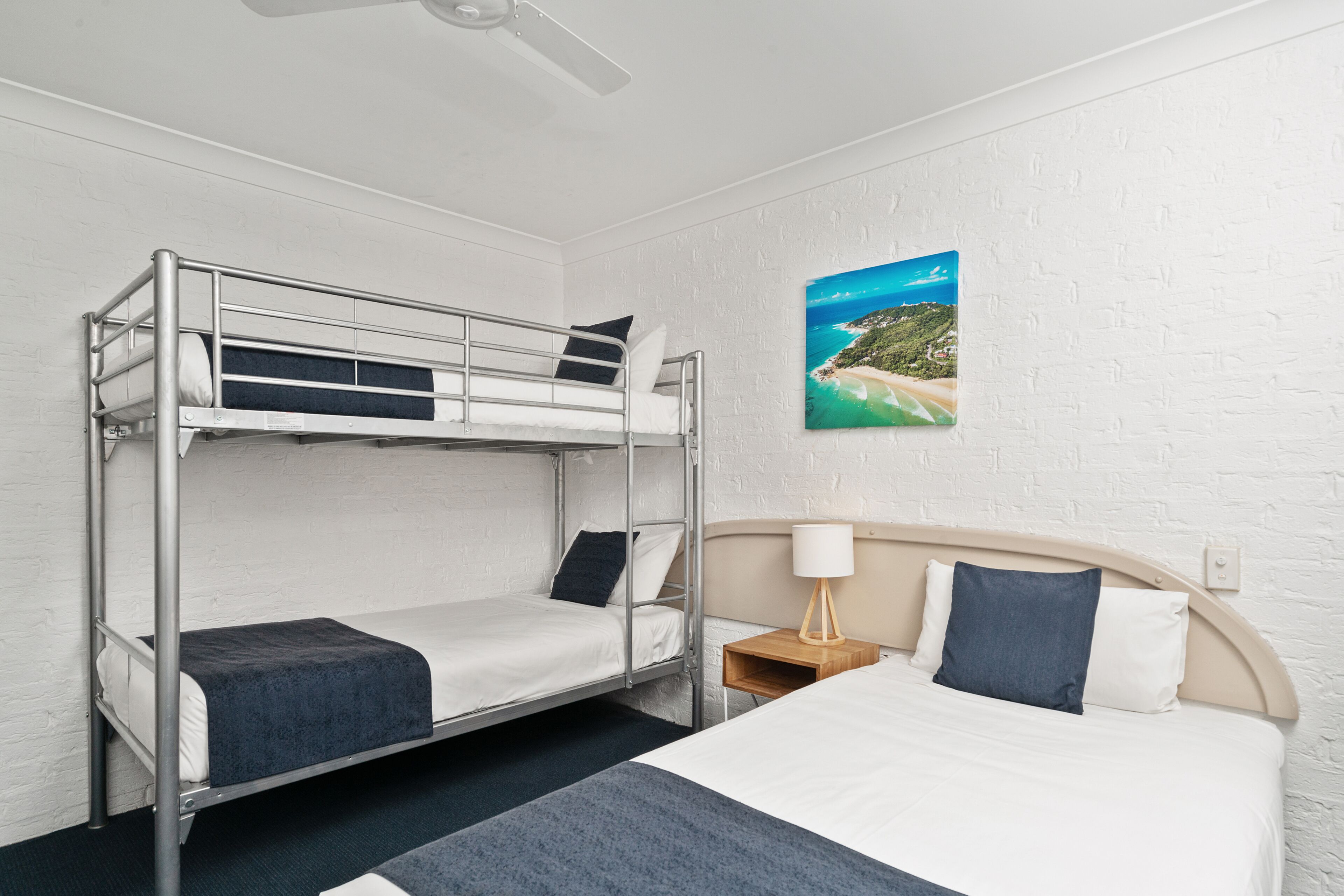 A hotel room with a bunk bed, a single bed, a nightstand with a lamp, and a wall-mounted painting.