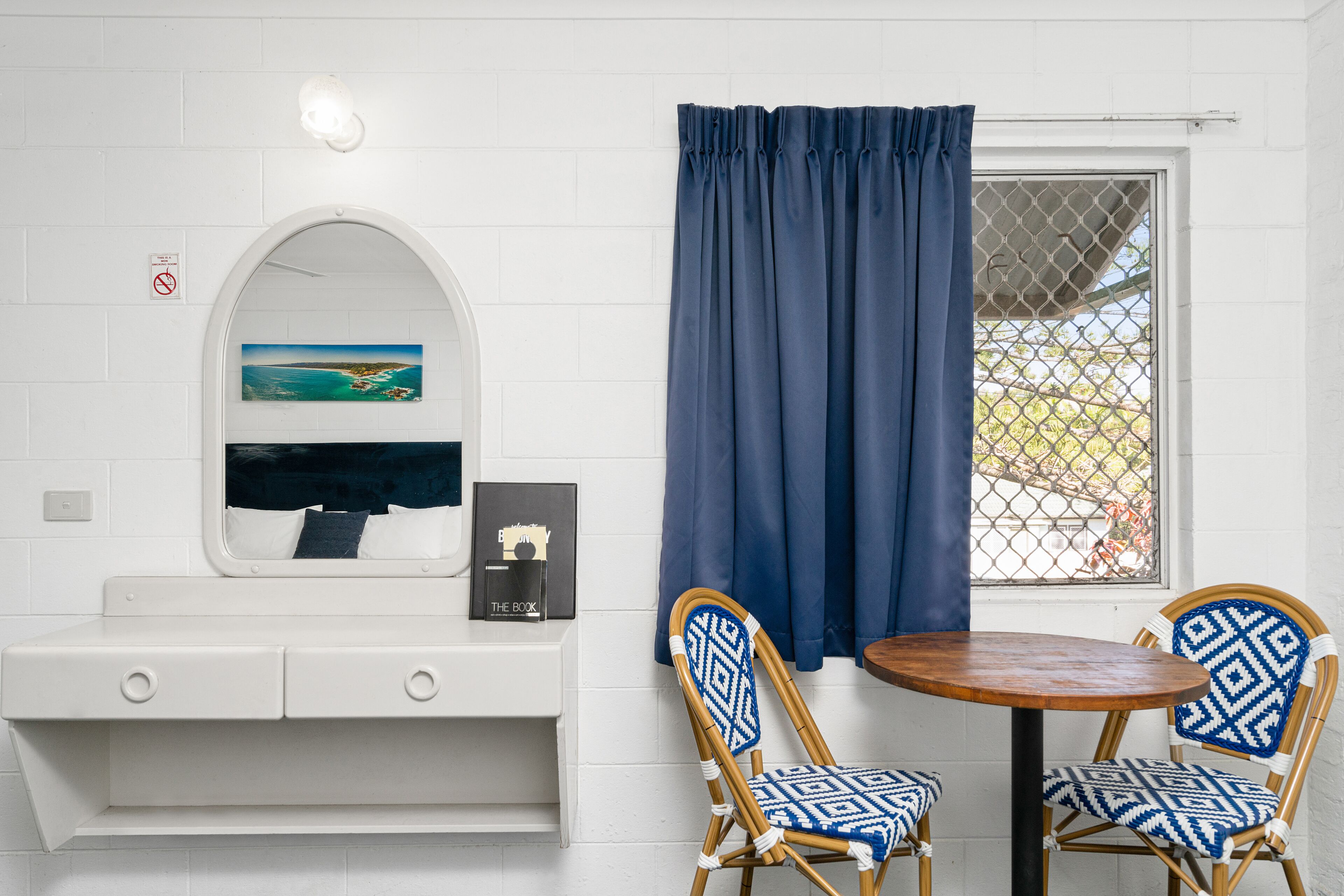 Una habitación con pared de ladrillo blanco, una consola blanca con un cuadro enmarcado y un libro, dos sillas estampadas alrededor de una mesa de madera, y una ventana con cortinas azules.