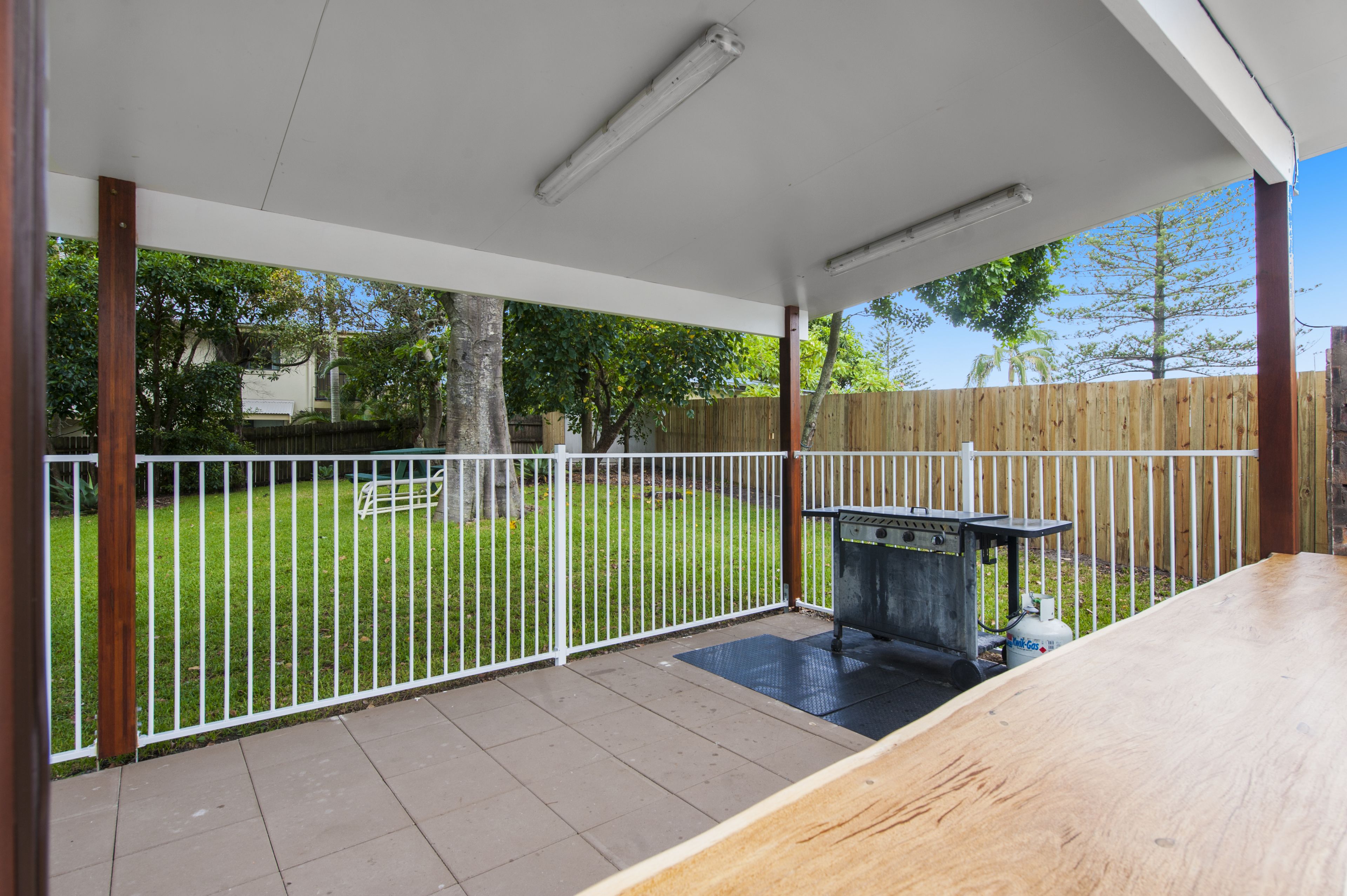 Ein überdachter Patio-Bereich mit Grill, weißer Brüstung und Blick auf einen grünen Garten.