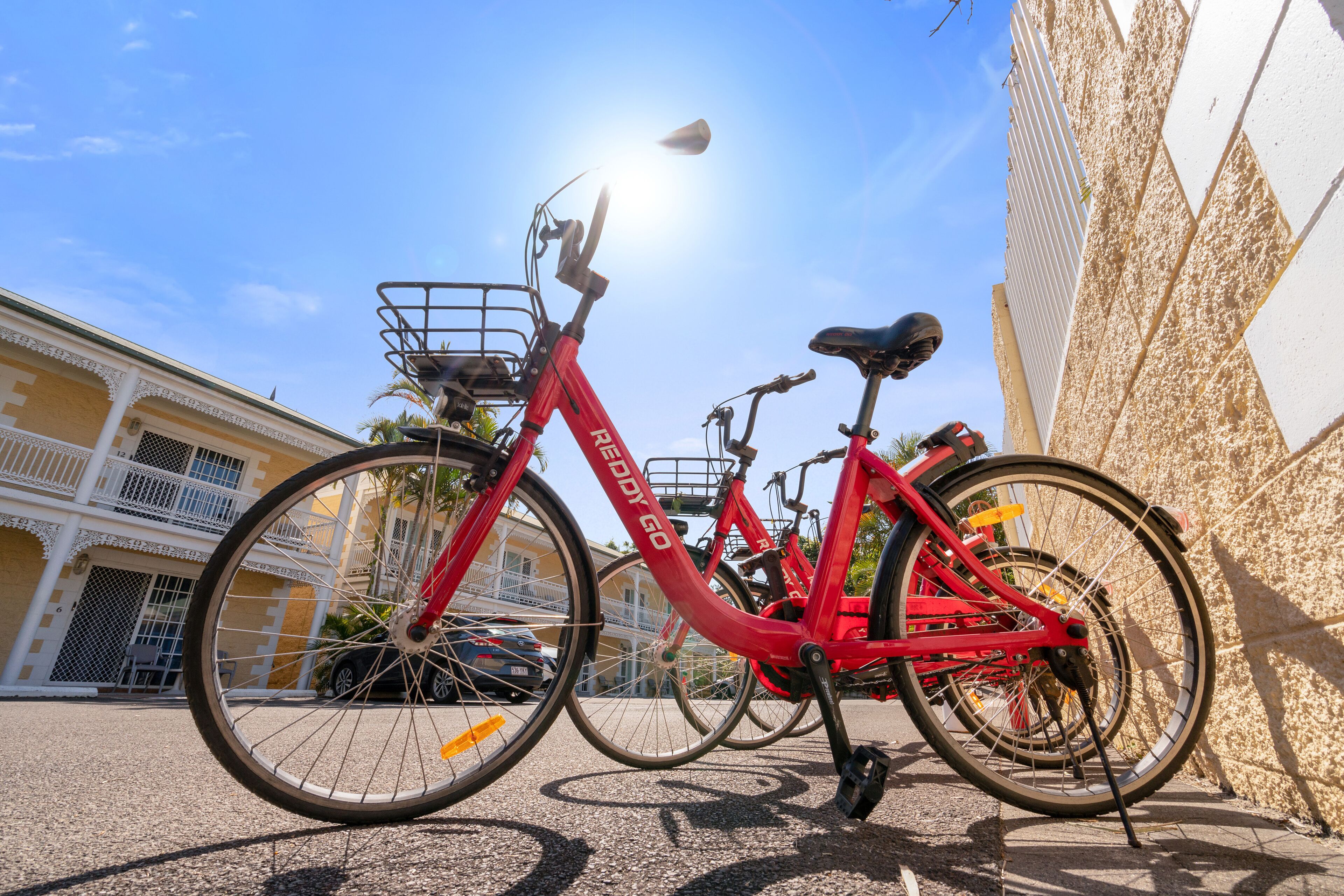 A red bicycle with a basket and a seat, parked against a wall with a building in the background.