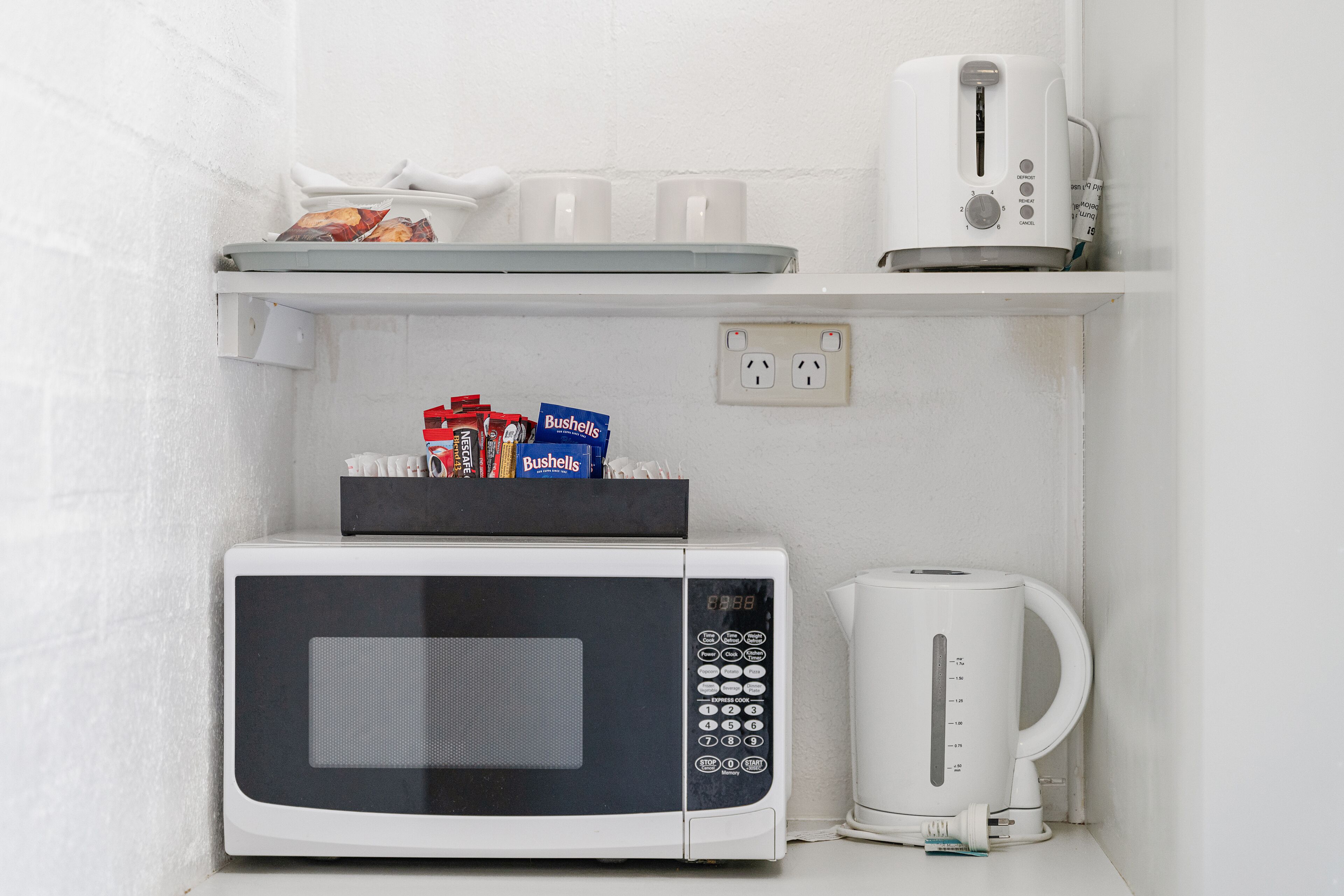 A compact kitchenette with a microwave, coffee maker, and a shelf with cups and snacks.
