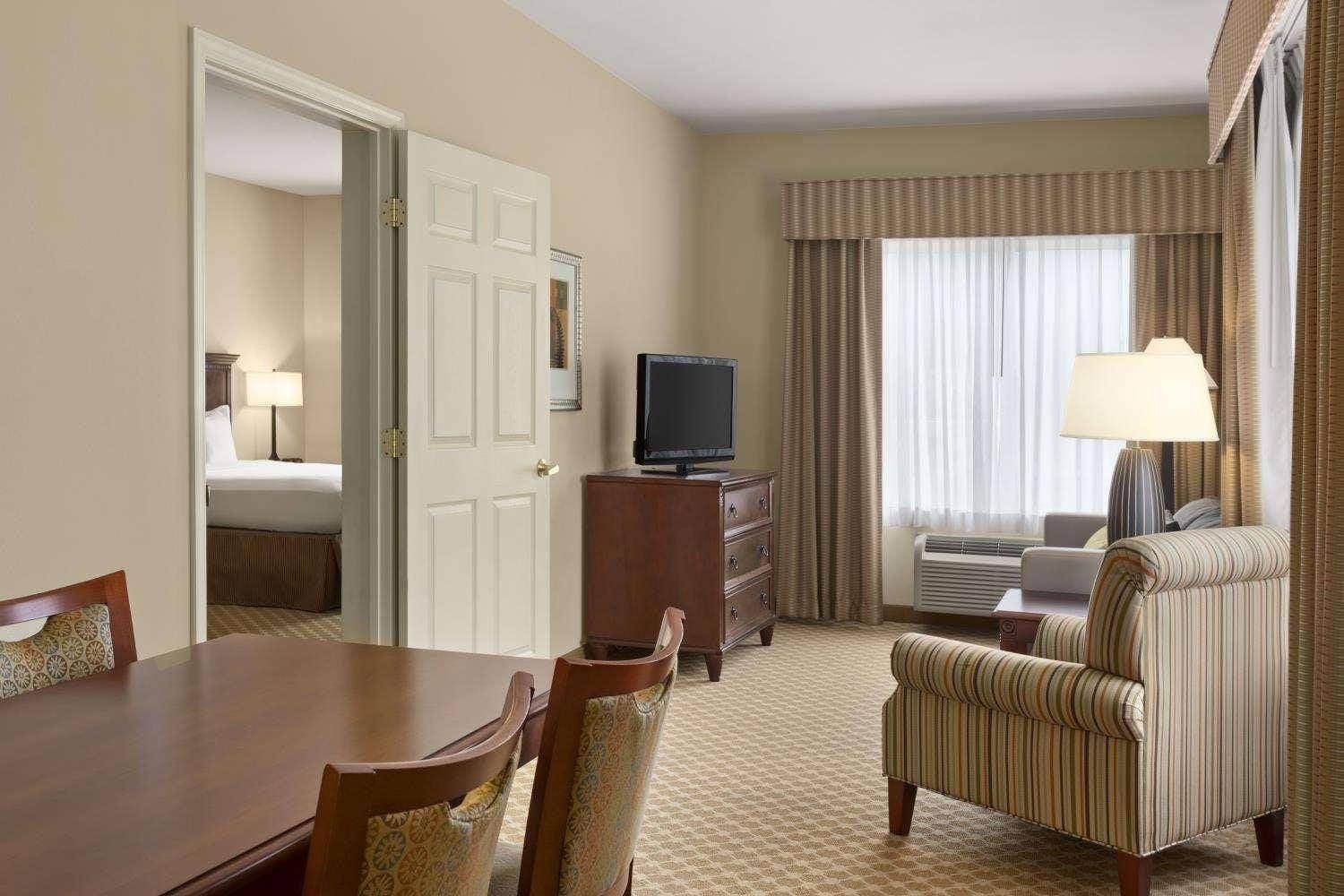 A hotel room with a bed, a television, a desk with a chair, a striped armchair, and a window with curtains.