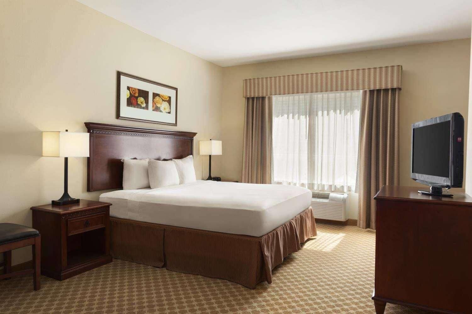 Habitación de hotel con una cama grande, dos mesitas de noche con lámparas, un televisor en una cómoda, una ventana con cortinas y un cuadro en la pared.