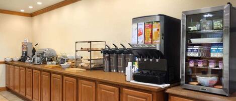Um balcão de pequeno-almoço self-service com máquinas de café, dispensador de bebidas e contentores refrigerados.