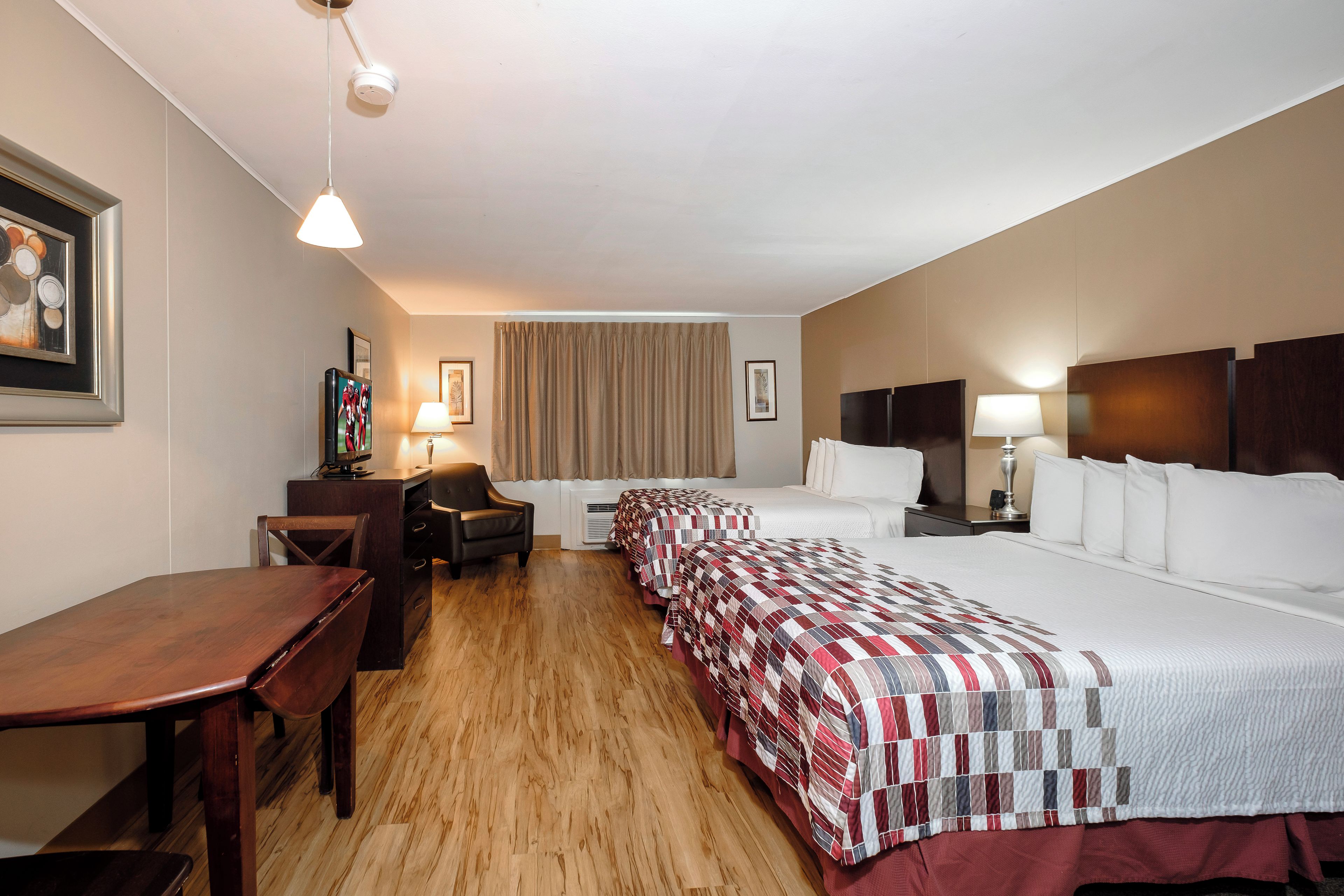 A hotel room with a wooden floor, a desk, two beds with red and white bedding, a chair, a TV, and a ceiling light.