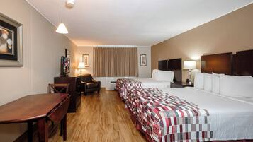 A hotel room with a wooden floor, a desk, two beds with red and white bedding, a chair, a TV, and a ceiling light.