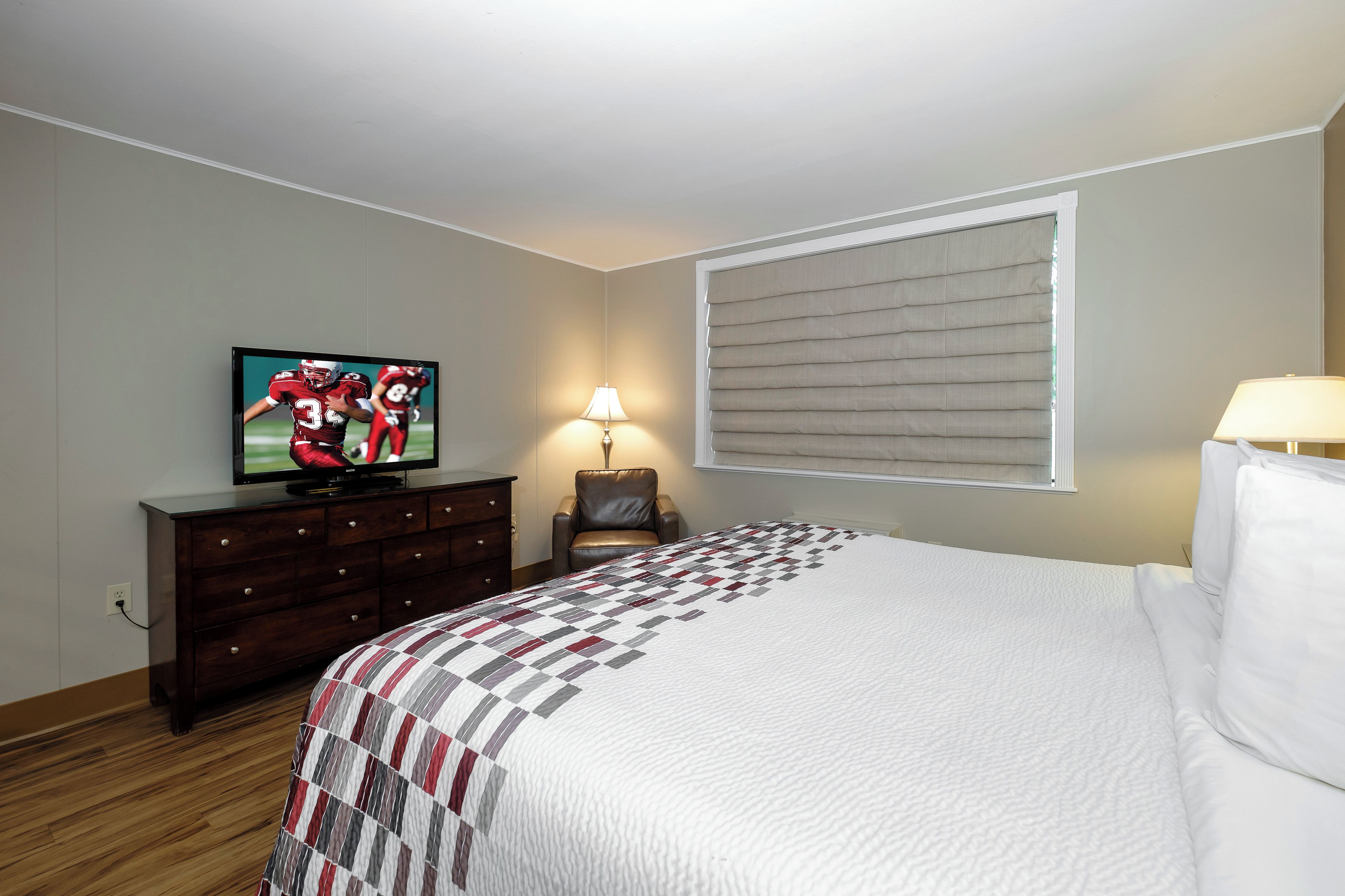 A hotel room with a bed, a television on a dresser, a lamp, and a window with blinds.