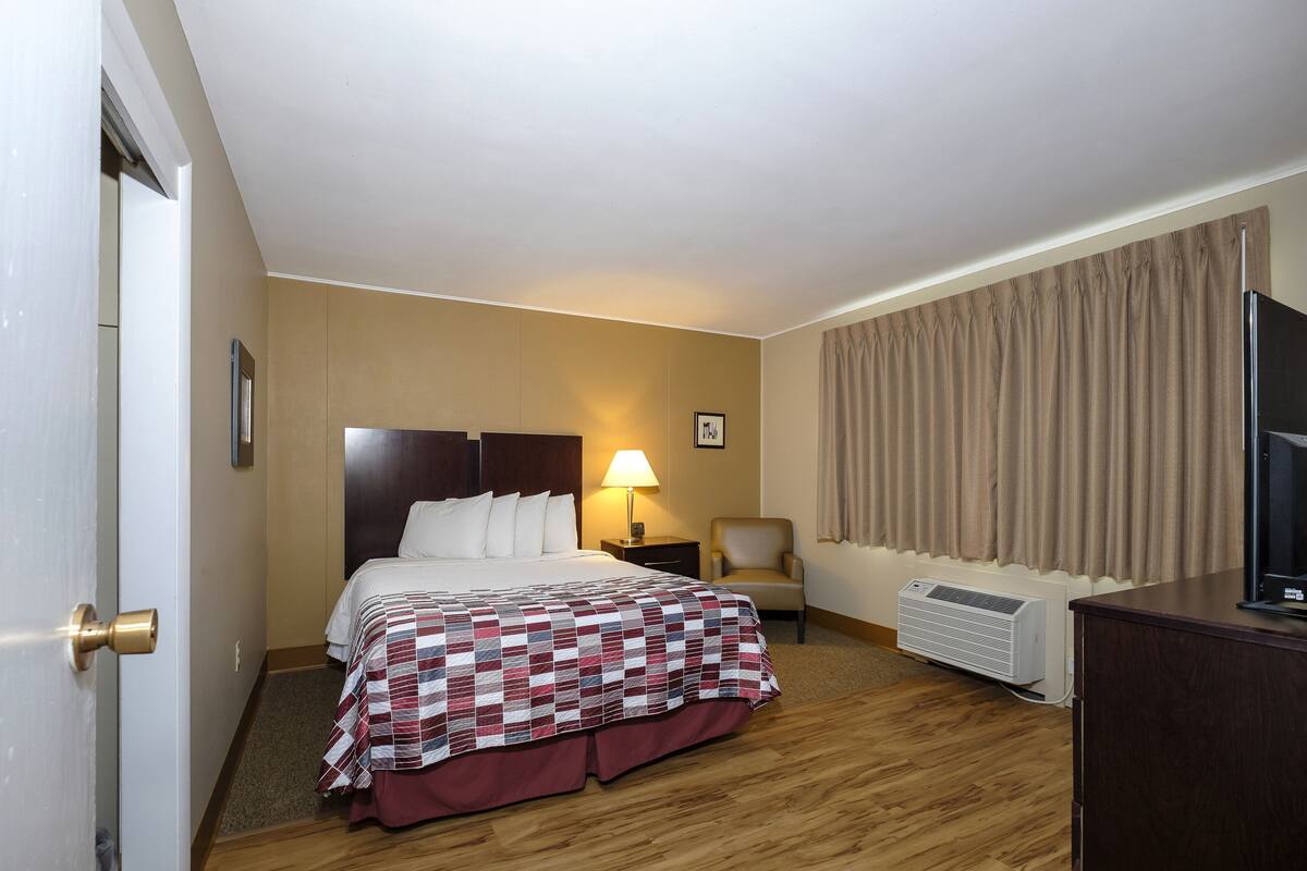 Quarto de hotel com cama, poltrona, escrivaninha e televisão.