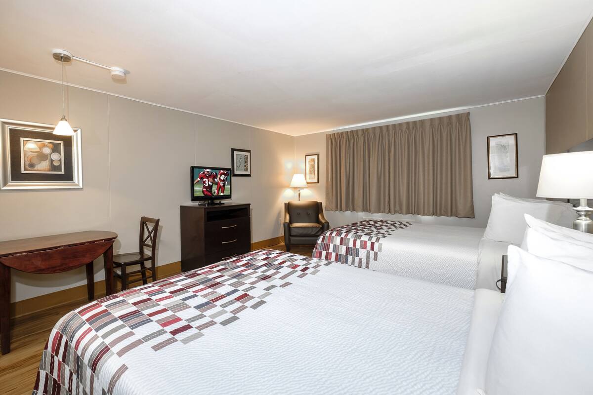 A hotel room with two beds, a desk, a chair, a TV, and a lamp.