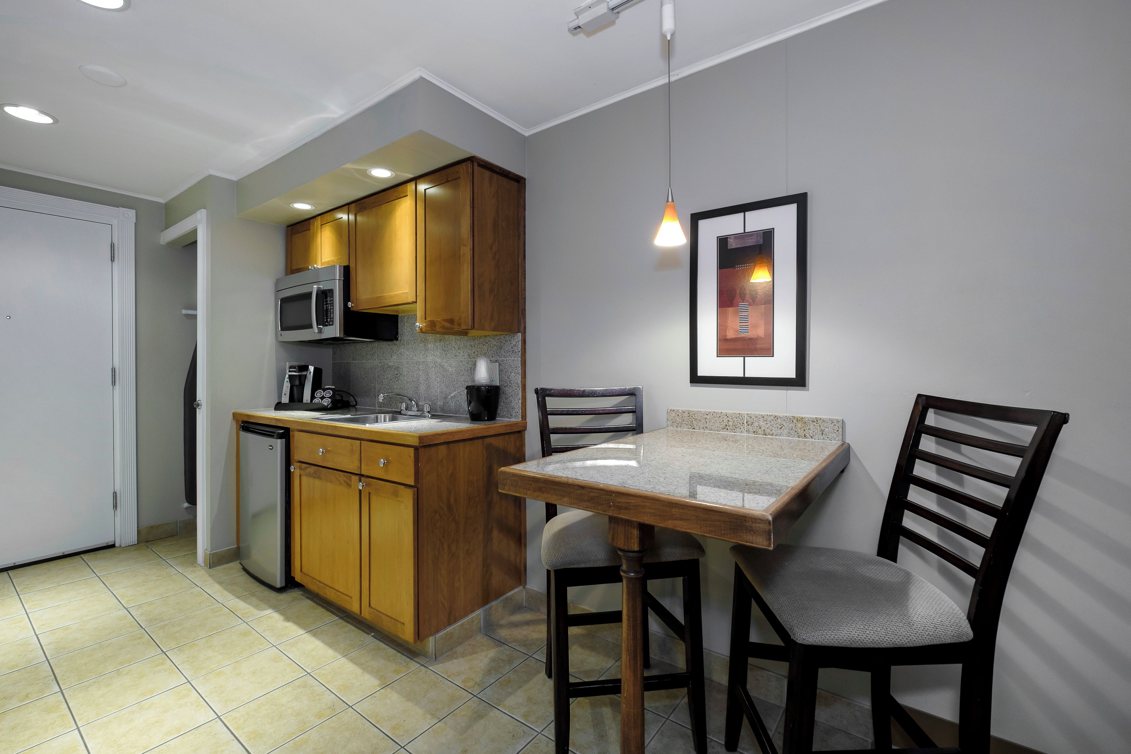 A compact kitchen with wooden cabinets, a microwave, a sink, and a small refrigerator. There is a dining table with two chairs and a pendant light above it.