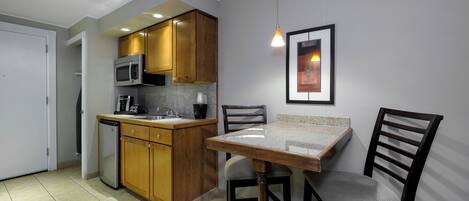 A compact kitchen with wooden cabinets, a microwave, a sink, and a small refrigerator. There is a dining table with two chairs and a pendant light above it.